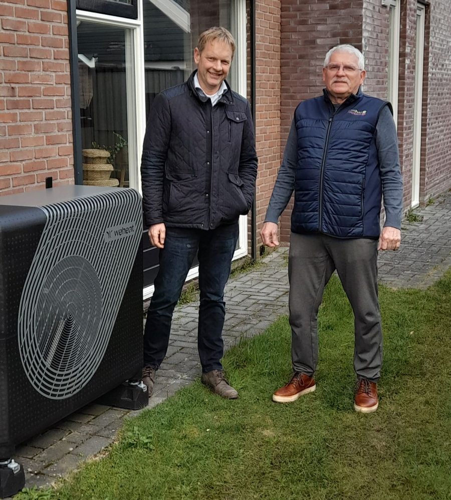 Full electric warmtepomp naast vrijstaande jaren 60-woning met twee mannen die verduurzaming bespreken.
